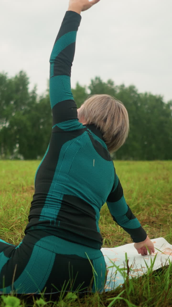 vista trasera de una mujer acostada en una alfombra de yoga, practicando la postura de flexión lateral con los brazos extendidos, y sentada en una postura lograda bajo un cielo nublado, en un vasto campo de hierba con árboles en la distancia