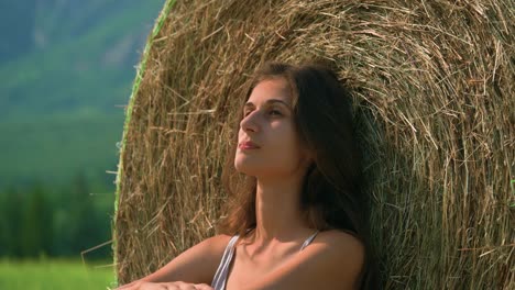 a beautiful brunette woman sitting calmly in thought against a large hay bale with a straw of grass in her mouth as she ponders