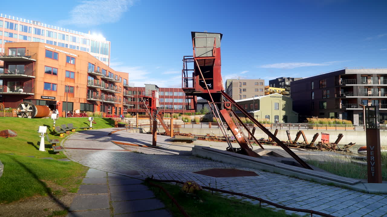 Historic shipyard cranes stand preserved beside new apartment buildings in Tromsø’s redeveloped Vervet harbour district on a bright sunny day. Foot traffic and cafes nearby