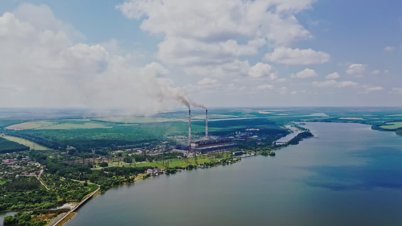 Aerial view of plant on the bank of the river in summer. View from above on natural background of beautiful river and factory with smoke from pipes. Slow motion. Camera moves up.
