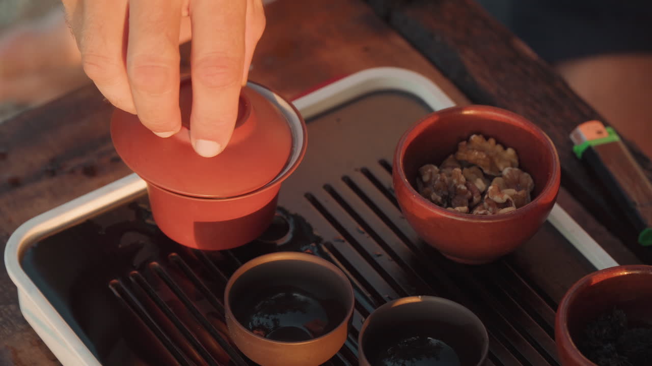 Beach tea ceremony with clay gaiwan, couple sharing quiet moment as hand lifts lid over steaming cup on wooden tray, sand and ocean blurred in background, warm golden light, slow intimate ritual
