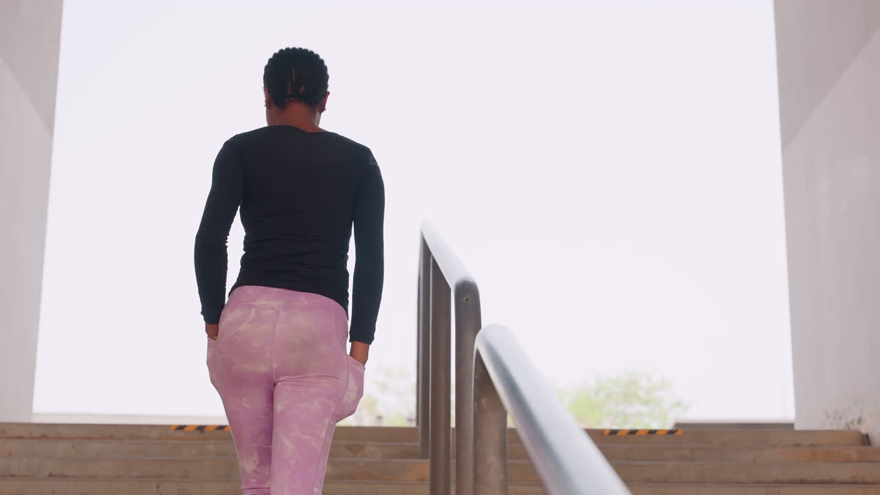black woman climbing outdoor stairs, black top pink leggings, confident stride ascending concrete stairwell, back view, soft morning light, urban commuter energy, fitness warmup, student transition