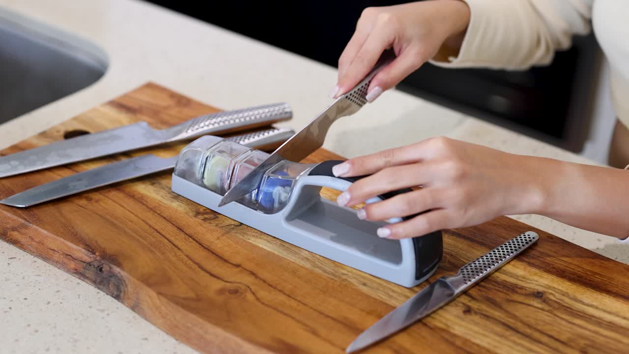 A person sharpens a knife using a handheld sharpener on a wooden board in a well-lit kitchen
