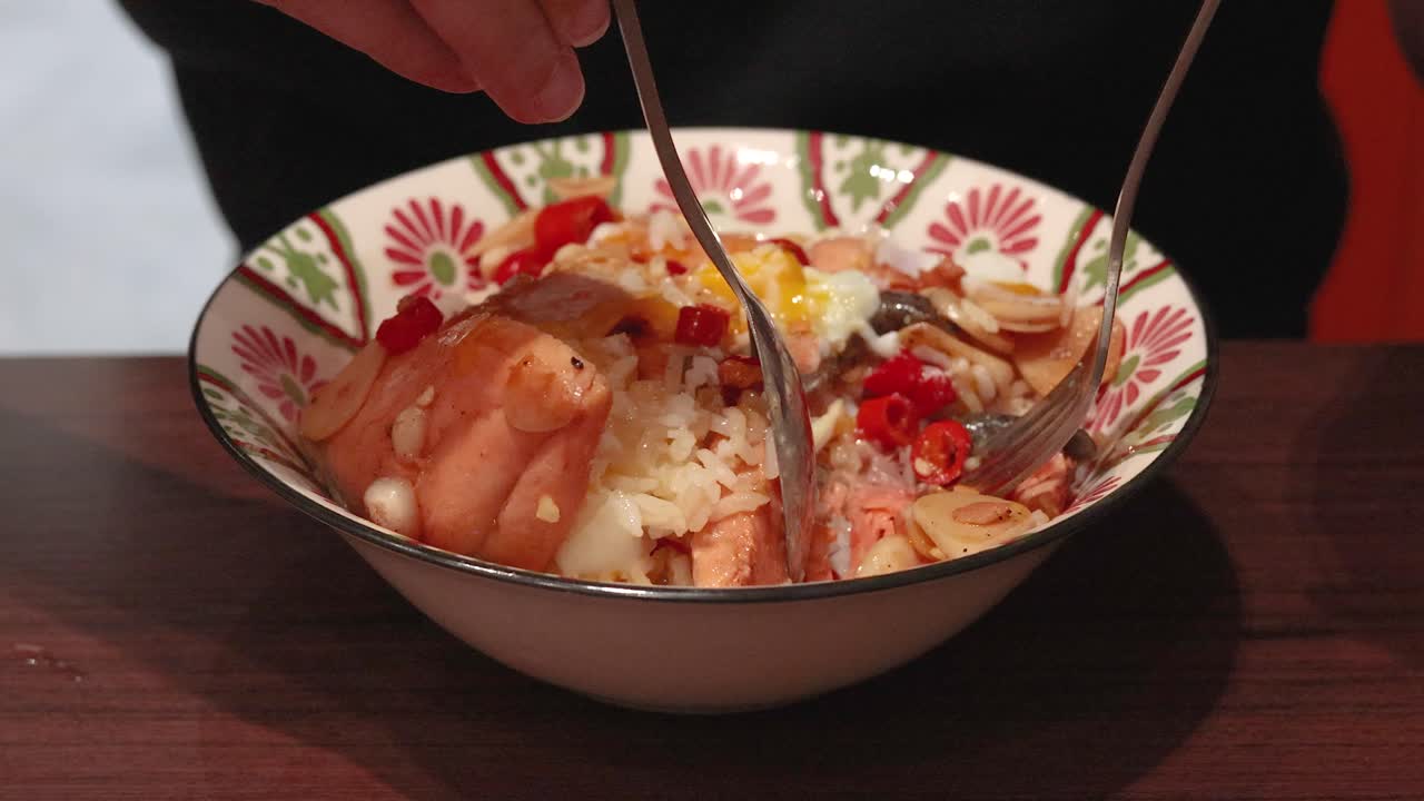 Hands use spoon and fork to mix cooked salmon, rice, garlic, and chili in a patterned bowl under warm indoor lighting, viewed from a close-up perspective