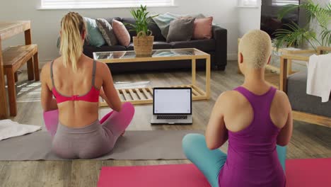 Diverse-female-couple-meditating-and-practicing-yoga-with-laptop-in-living-room