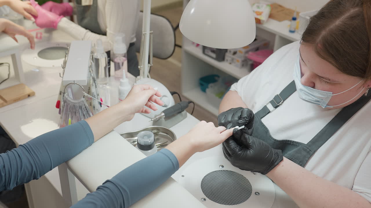 Two nail technicians focused on filing client nails with precision, surrounded by organized manicure tools and bottles on white salon table under bright task lamps in clean indoor workspace setting