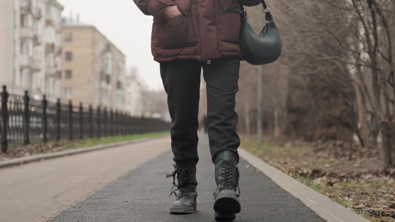 Gros plan sur des bottes marchant sur un chemin goudronné, un navetteur solitaire portant un manteau doudoune marron et un sac à bandoulière, rue résidentielle avec clôture et arbres dénudés, lumière hivernale tamisée, démarche régulière et délibérée.
