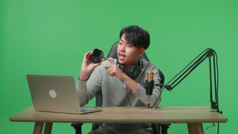 a smiling asian man with headphone and computer reviewing camera len while sitting in front of green screen background