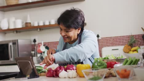 Asiatische-Seniorin-Nutzt-Digitales-Tablet,-Während-Sie-Zu-Hause-In-Der-Küche-Gemüse-Hackt