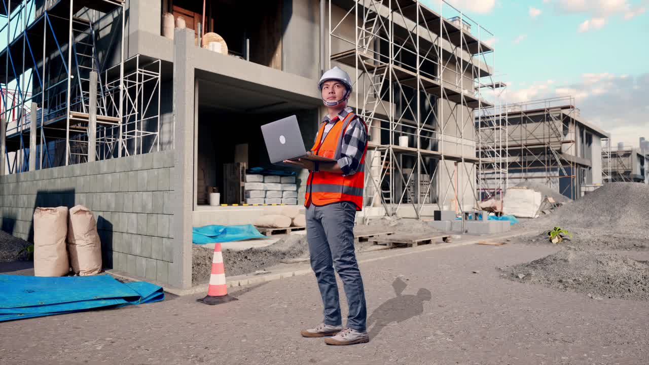 Full Body Side View Of Asian Male Engineer With Safety Helmet Working On A Laptop And Looking Around While Standing At Building & Construction Site in Progress to New House