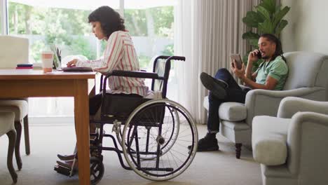Happy-biracial-woman-in-wheelchair-using-laptop,-with-male-partner-talking-on-phone-in-living-room