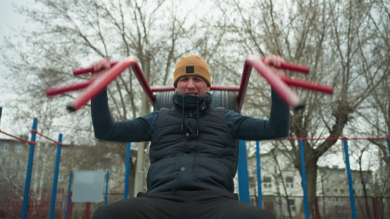 A coach wearing a brown beanie and black jacket is doing pull-ups on outdoor equipment, gripping red bars, in the background, a boy is running close to the coach