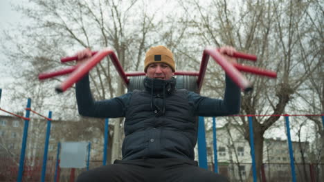 a coach wearing a brown beanie and black jacket is doing pull-ups on outdoor equipment, gripping red bars, in the background, a boy is running close to the coach