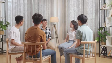 아시안 십대 그룹은 의자에 앉아서 집에서 논의하는 원을 형성하고 있습니다. 모두가 카레드 셔츠를 입은 소년이 자신을 소개하는 것을 듣고 있습니다.