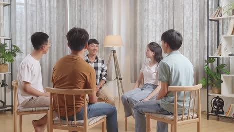 asian teen group sitting in chairs forming a circle discussing at home. everybody is listening to a boy in plaid shirt introducing himself