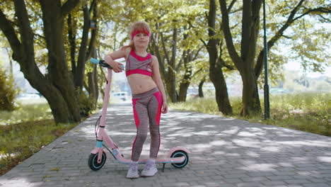 athletic fit young girl rides on electric scooter, standing near bike looking at camera in park