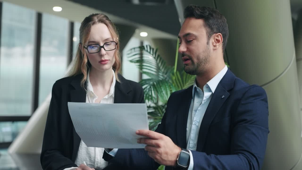 Amministratore delegato professionista fornisce un feedback dettagliato a una dipendente mentre esaminano insieme documenti cartacei e grafici di un progetto in un moderno ufficio aziendale