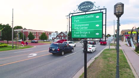 cars driving down a main street in owosso, mi, with a signage in the foreground.