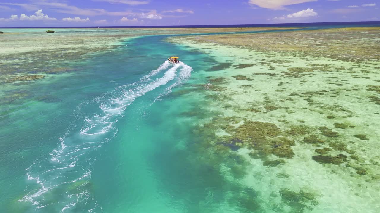 A stunning aerial shot captures a speedboat as it carves a path through a narrow, deep blue channel in the middle of a vast, shallow coral reef. The vibrant turquoise water contrasts beautifully