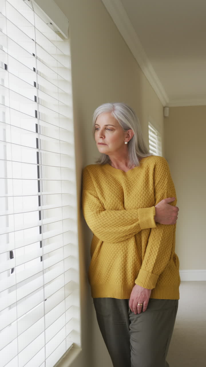 집에서 창밖을 바라보는 사려 깊은 백인 고령 여성의 수직 비디오