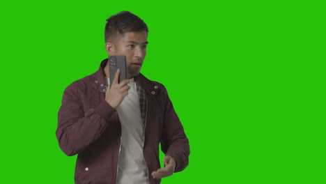 studio shot of casually dressed smiling young man taking call on mobile phone against green screen 2
