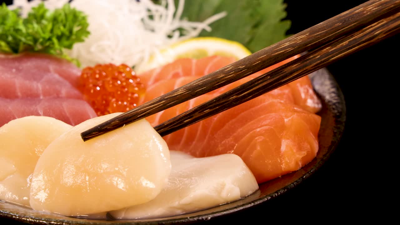 Chopsticks delicately pick up a scallop sashimi from a vibrant seafood platter with salmon, tuna, roe, and garnishes under bright studio lighting