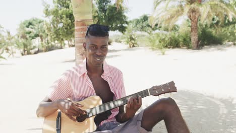 Retrato-De-Un-Hombre-Afroamericano-Feliz-Tocando-La-Guitarra-Y-Cantando-En-Una-Playa-Soleada,-Cámara-Lenta