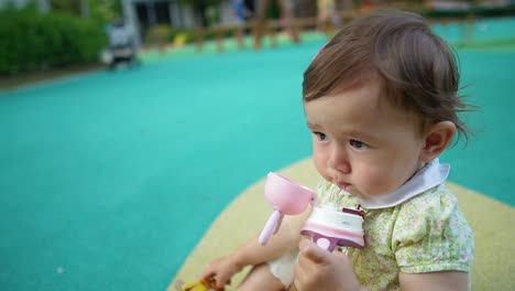 Una-Dulce-Niña-De-Un-Año-Está-Sentada-En-El-Suelo-Del-Patio-De-Recreo-Sosteniendo-La-Botella-En-Las-Manos,-Bebiendo-Agua,-Espacio-De-Copia-De-Plantilla-En-Cámara-Lenta