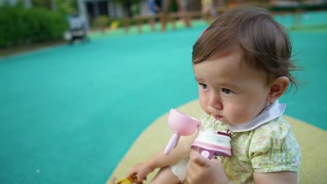 a sweet one-year-old girl is sitting on playground floor holding the bottle in hands, drinking water, template slow-motion copy space