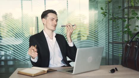 young, stylish man in business attire is sitting at a desk in a modern office, speaking through voice messages on his smartphone with a laptop in front of him