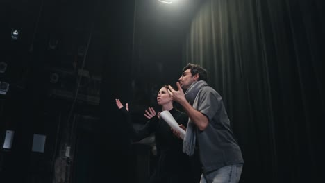 A-confident-male-brunette-director-with-glasses-in-a-T-shirt-and-a-scarf-walks-to-the-last-stage-of-the-theater-with-gray-curtains-together-with-his-student-actress-in-a-black-suit-and-expressively-play-their-roles