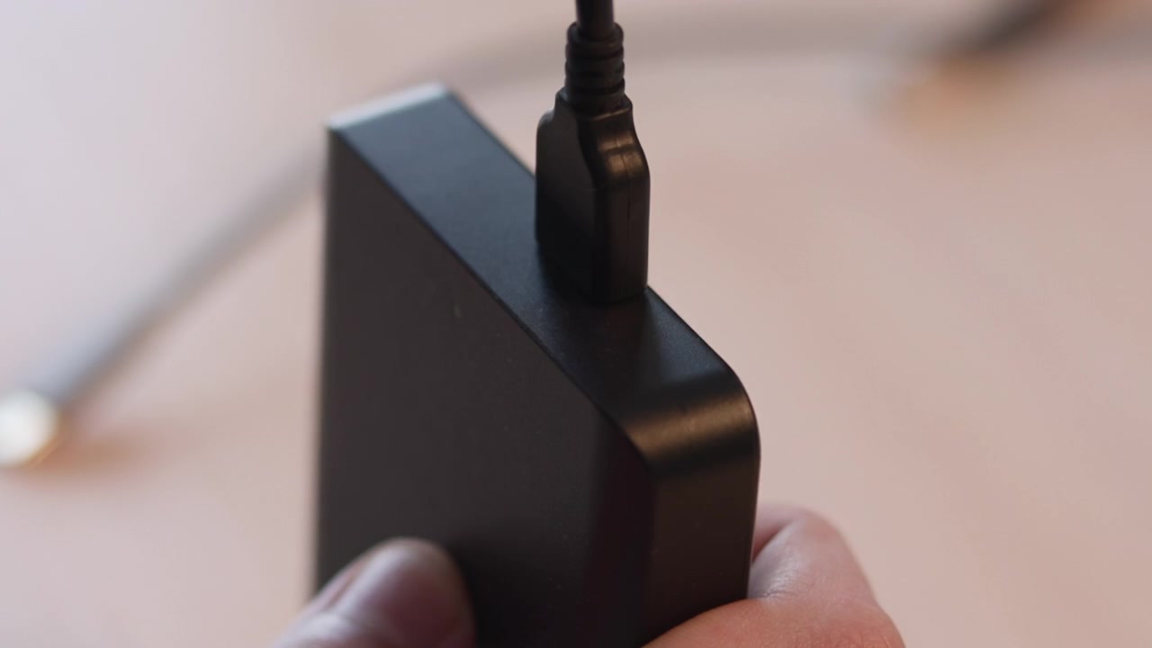 Scène d'intérieur en gros plan montrant une main stabilisant un adaptateur noir mat tandis qu'un câble micro USB s'insère dans le port supérieur encastré, avec un flou doux et chaud, un détail de matériel compact et une connexion clairement visible.
