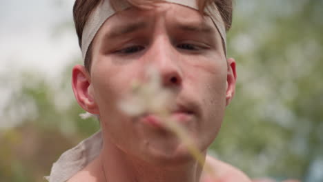 close-up of young adventurer blowing dandelion flower with intense, rough expression while standing outdoors surrounded by greenery under daylight sky, showing determination and emotion