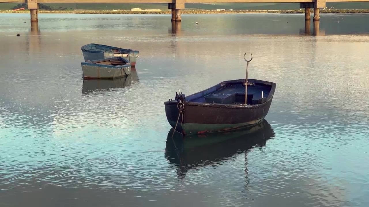 Gamle træfiskebåde flydende på det rolige vand ved Barbate, Andalusien, Spanien, der reflekterer himlen og broen i en fredfyldt, malerisk scene.