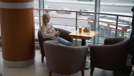 young lady seated alone in mall holding phone, gazing out large window with busy city view, cars passing by on snowy streets as she enjoys peaceful moment with coffee on wooden table