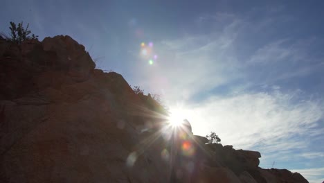 Looking-up-pan-shot-of-sunshine-dazzling-behind-the-mountain-rock-silhouette-during-sunny-day-with-blue-sky
