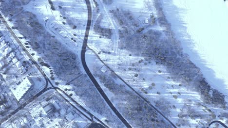 Panorámica-Aérea-A-Vista-De-Pájaro-A-La-Izquierda-De-Una-Autopista-Y-Un-Río-Cubierto-De-Nieve-Helada-Junto-Al-Parque,-Al-Lado-De-La-Ciudad