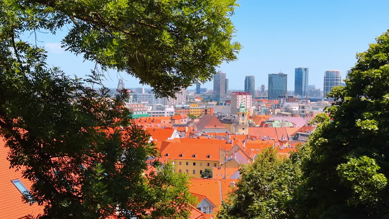 Orange roofs of the old town can be seen through the green tree branches. Modern high-rises at backdrop. Bratislava, Slovakia