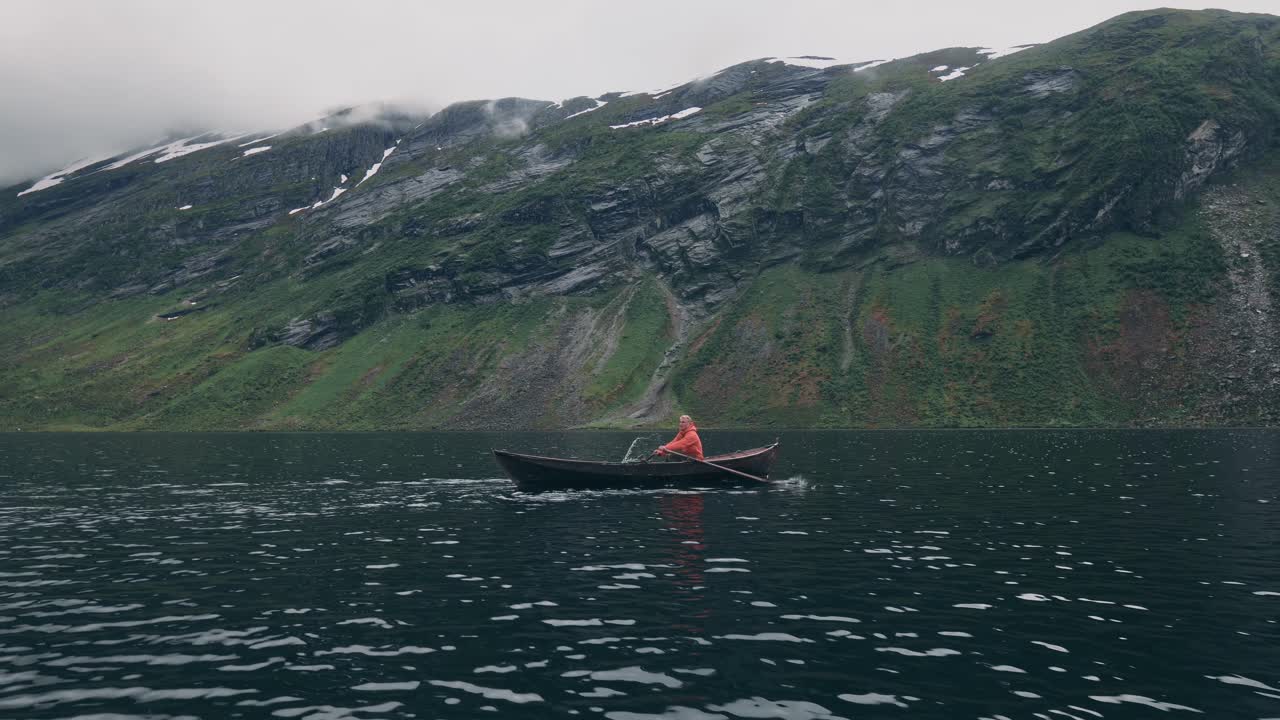 Person rowing a small rowboat in the middle of a large lake, mountains in background.