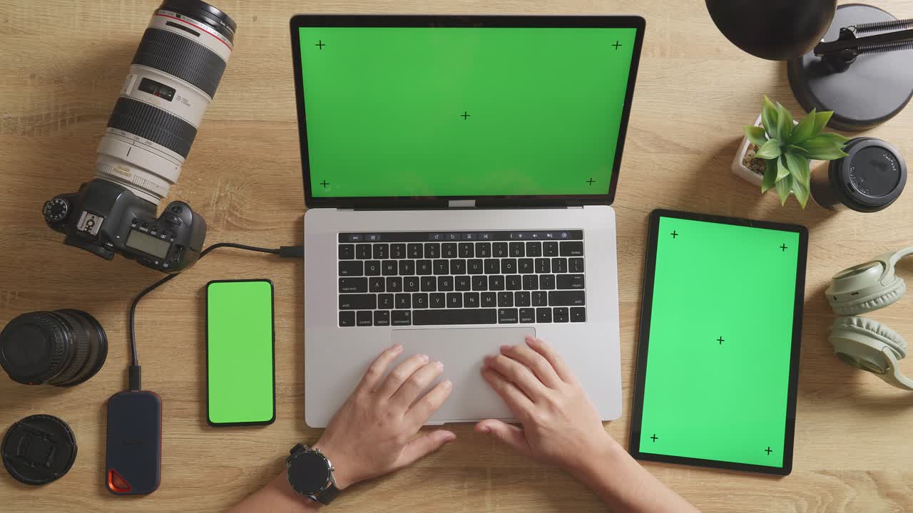 vista superior de cerca de las manos del editor de video masculino usando una computadora portátil de pantalla verde con un teléfono inteligente y una tableta de pantalla verde junto a la cámara en el espacio de trabajo en casa
