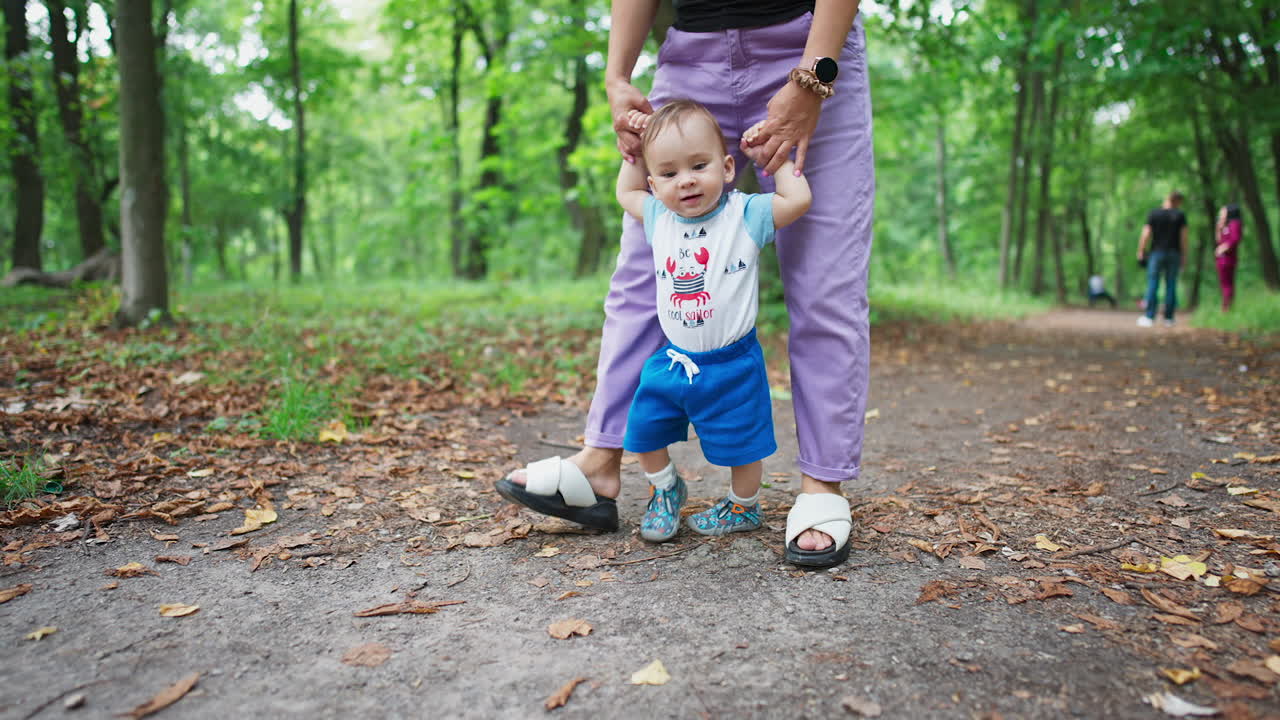 Teaching baby boy to walk. Mother is holding her baby by the hands and leading her child along the path in the park. Nature blurred backdrop.