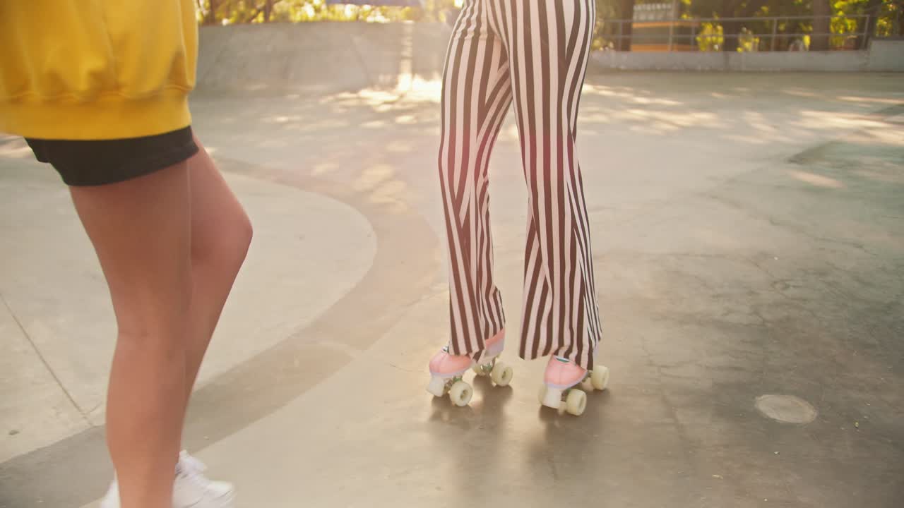 una chica morena con una camiseta amarilla ayuda a una chica con un corte de pelo corto en una camiseta púrpura y auriculares rojos con pantalones a rayas a patinar en un parque de patinaje en verano