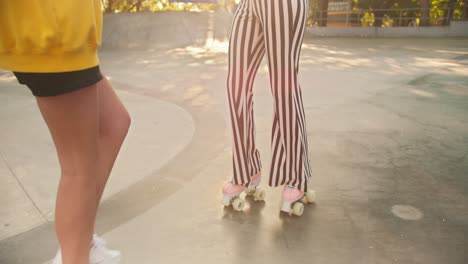 A-brunette-girl-in-a-yellow-t-shirt-helps-a-girl-with-a-short-haircut-in-a-purple-top-and-red-headphones-with-striped-pants-ride-roller-skates-in-a-skate-park-in-summer