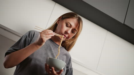 woman wearing casual outfit bends slightly while eating pasta with chopstick from bowl in modern kitchen, focused expression, strands of long pasta hanging from chopstick
