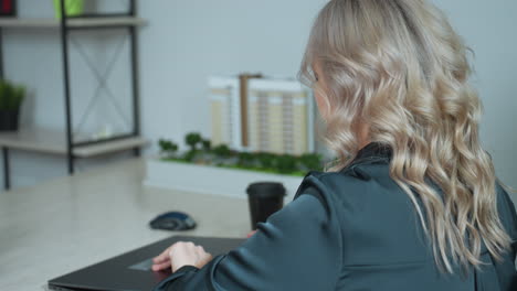 rear view of professional woman at desk adjusting phone and file while opening laptop in organized office with scale model and modern decor, preparing for productive day