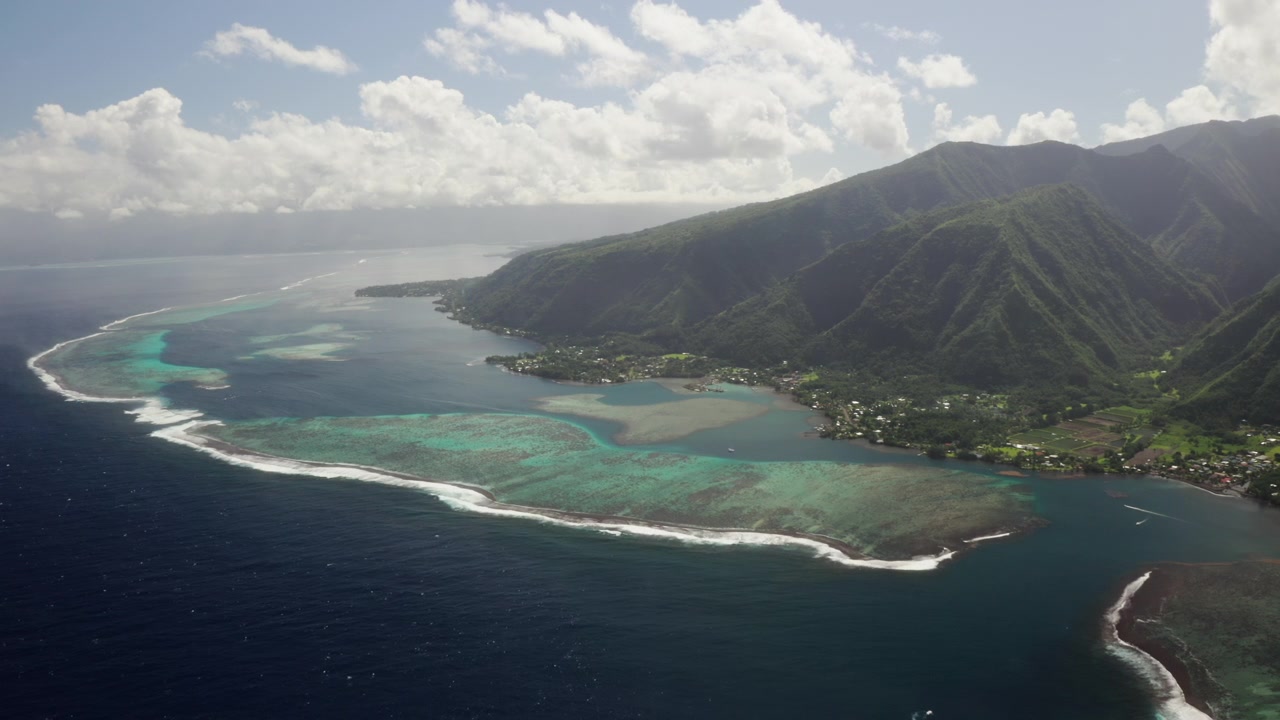 Un drone aereo si libra su un canale della barriera corallina di Teahupoo, Tahiti, con sagome di montagne che svaniscono sotto nuvole pesanti mentre la marea si muove attraverso il corallo