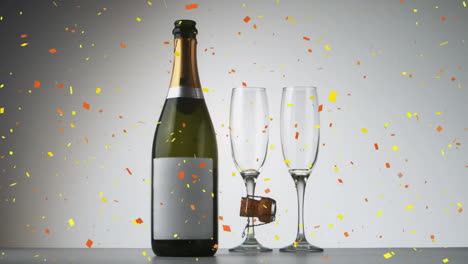 confeti cayendo sobre una botella de champán y dos vasos de champán contra un fondo gris
