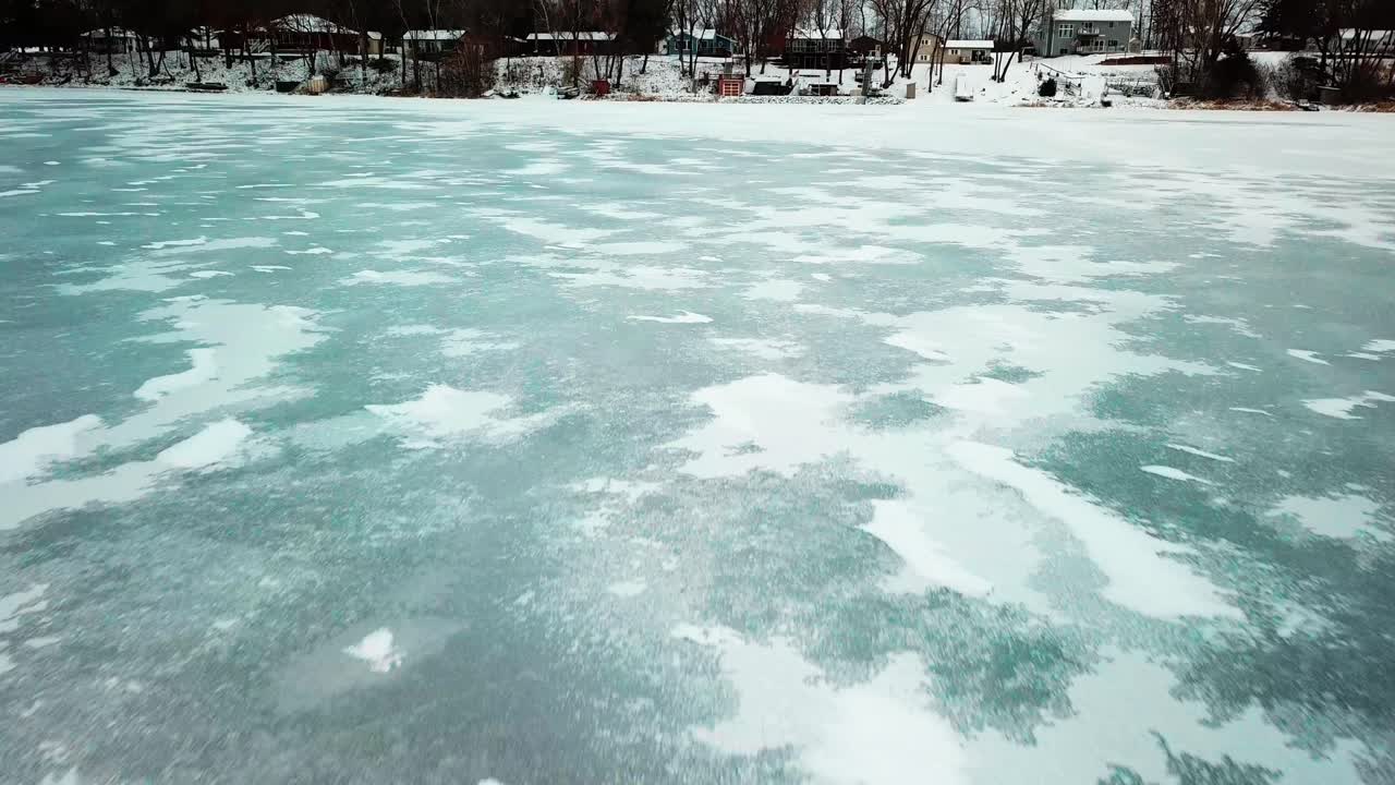 Low fly over ice on frozen lake and tilt up to reveal lakeside cabins during winter. 4K