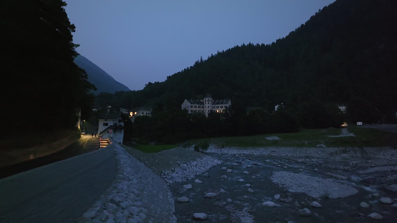A serene evening view of the Maloja Pass village in the Swiss Alps. An illuminated path runs alongside a rocky riverbed, with mountains silhouetted against the dusk sky, Switzerland