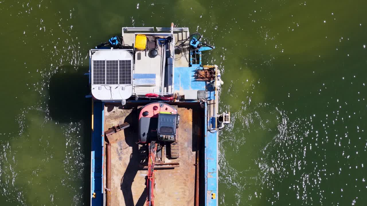 Top-down drone shot of industrial barge with excavator, solar panels, and engine on green water